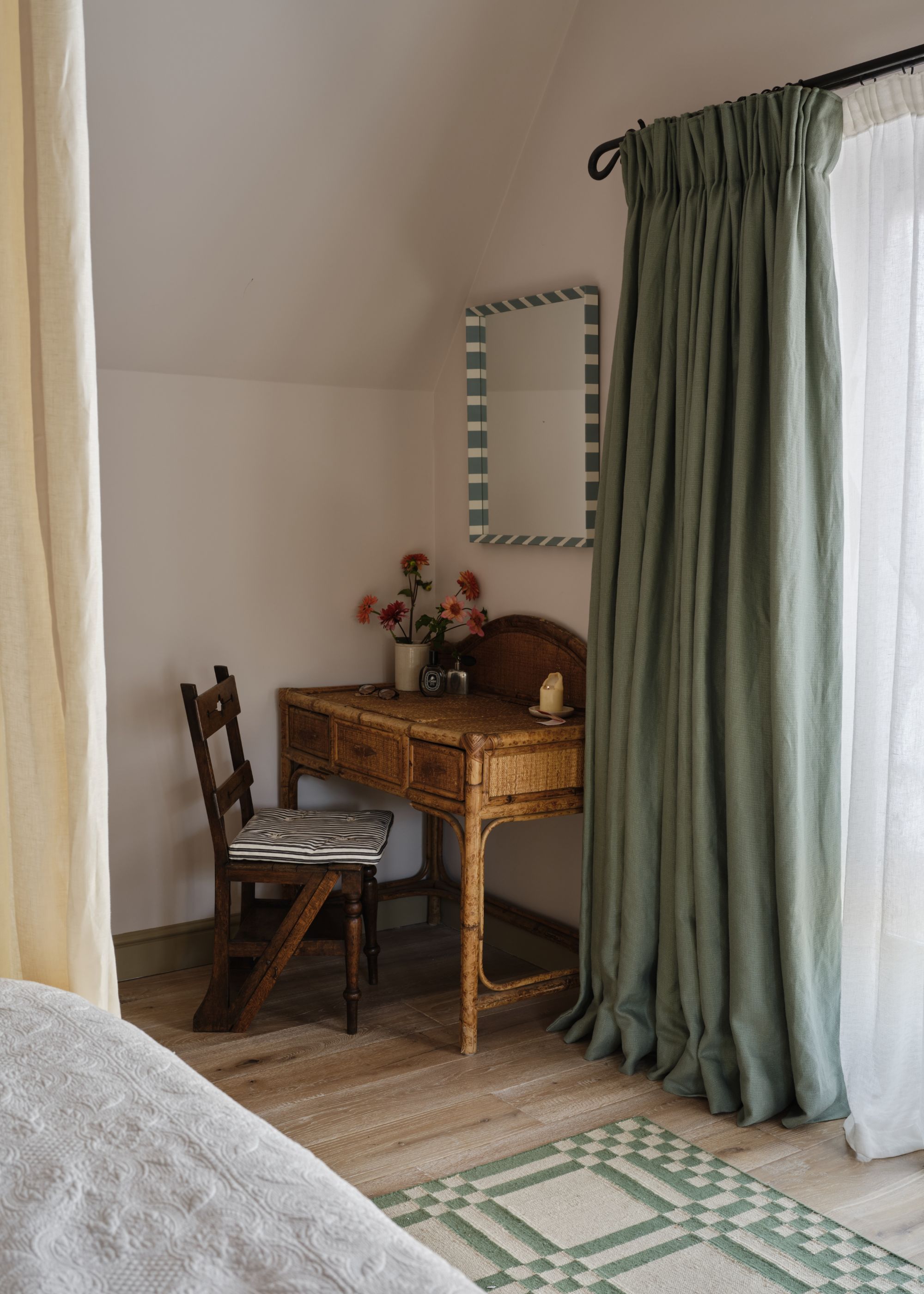 A neutral bedroom with soft white walls, wood floors, sage green curtains, a vintage wood desk with a chair, and a mirror on the wall.