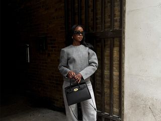 coco wears gray mono chrome outfit while holding a black handbag and wearing black pointed heels.