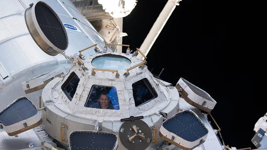 exterior view of a many-windowed module of the international space station, showing a smiling male astronaut at one of the windows