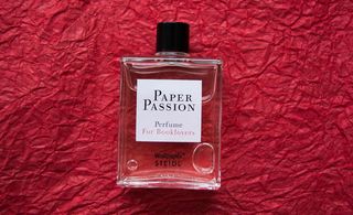 A square perfume bottle with the words "Paper Passion", lying on a piece of crumpled, red tissue paper