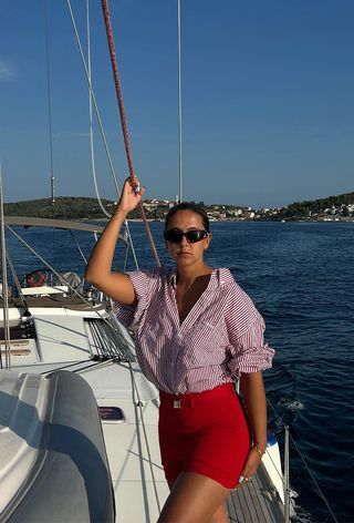 a photo showing what to wear on a boat with a woman wearing black sporty sunglasses with a red and white button-down shirt and red mini skirt