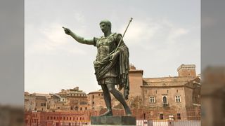A statue of Roman emperor Augustus. He has short hair and is wearing a decorated breast plate with his cape draping around from his back, around his waist, and then over his left arm. He has one pointed finger in the air and in his other hand he is holding a rod.