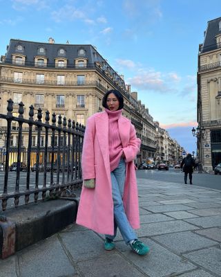 Woman in pink coat and sweater with light-wash jeans and green Adidas sneakers in Paris