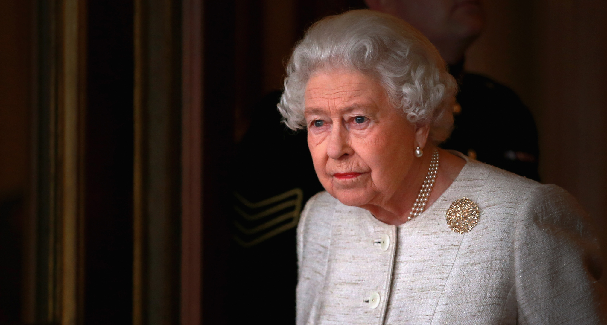 Queen Elizabeth wearing a white tweed jacket and brooch looking out a window