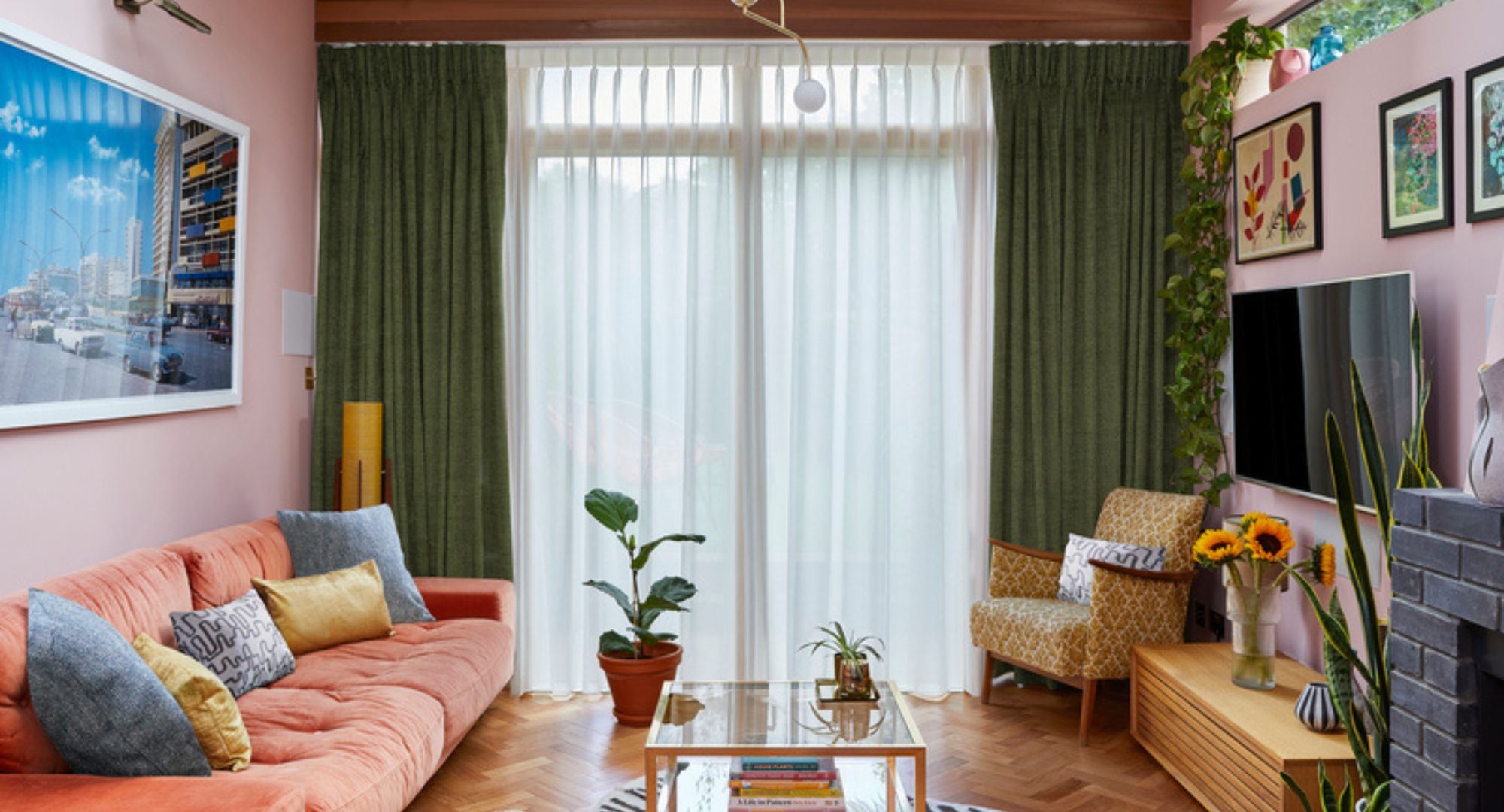 Pink living room with orange couch, plant pot, glass coffee table, pictures on the walls, wooden flooring and green curtains with sheer voile beneath