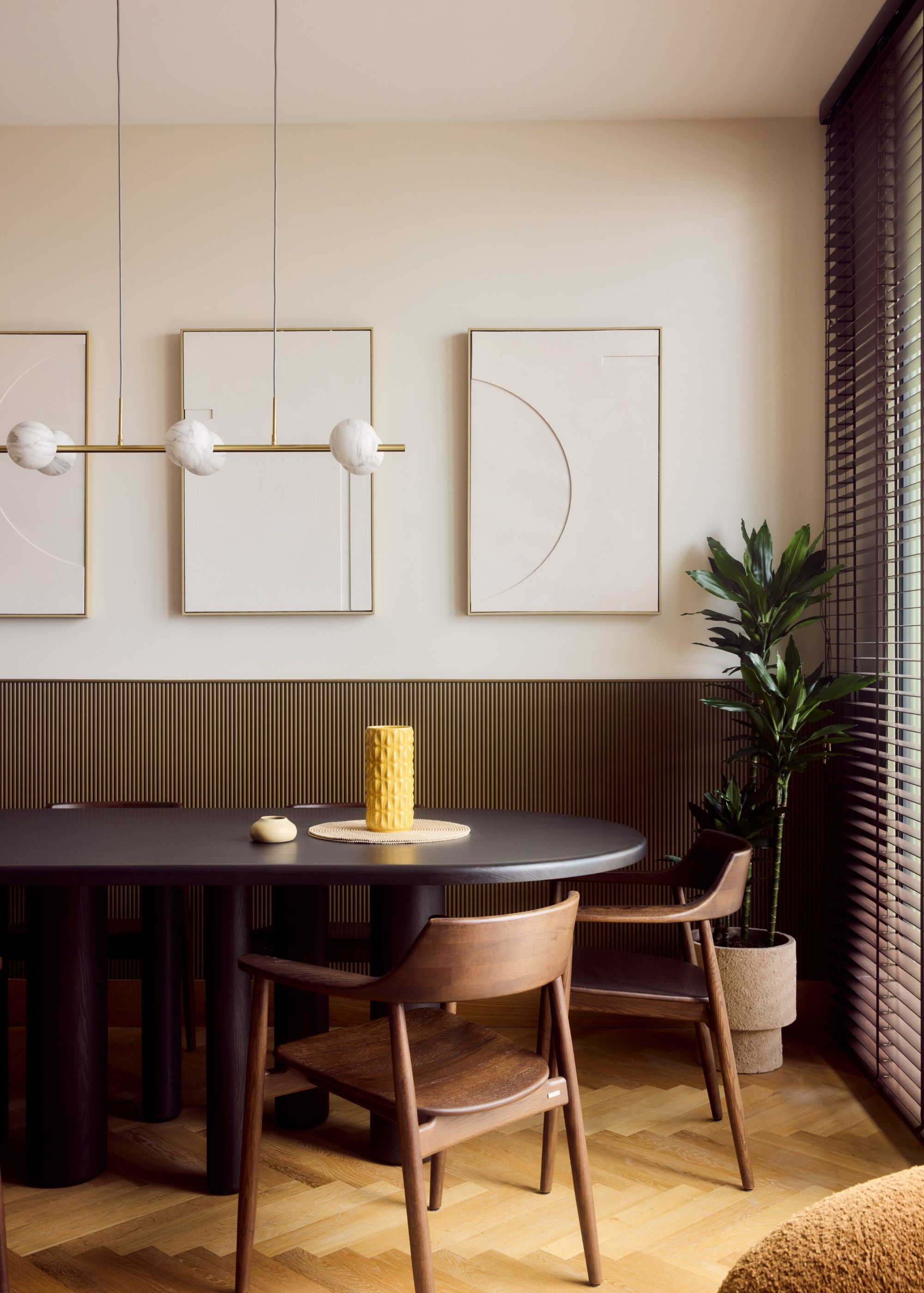Modern dining room with off-white walls, dark bronze wall panelling, a black modern dining table with two wooden chairs, parquet flooring and three white modern artworks.