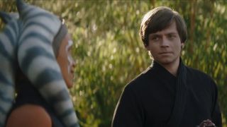 Still from the Star Wars T.V. show The Book of Boba Fett. Ahsoka (orange face with white markings and blue and white stripped head tails) and Luke Skywalker (white male, short floppy brown hair wearing dark Jedi robes) are having a chat by some green trees.