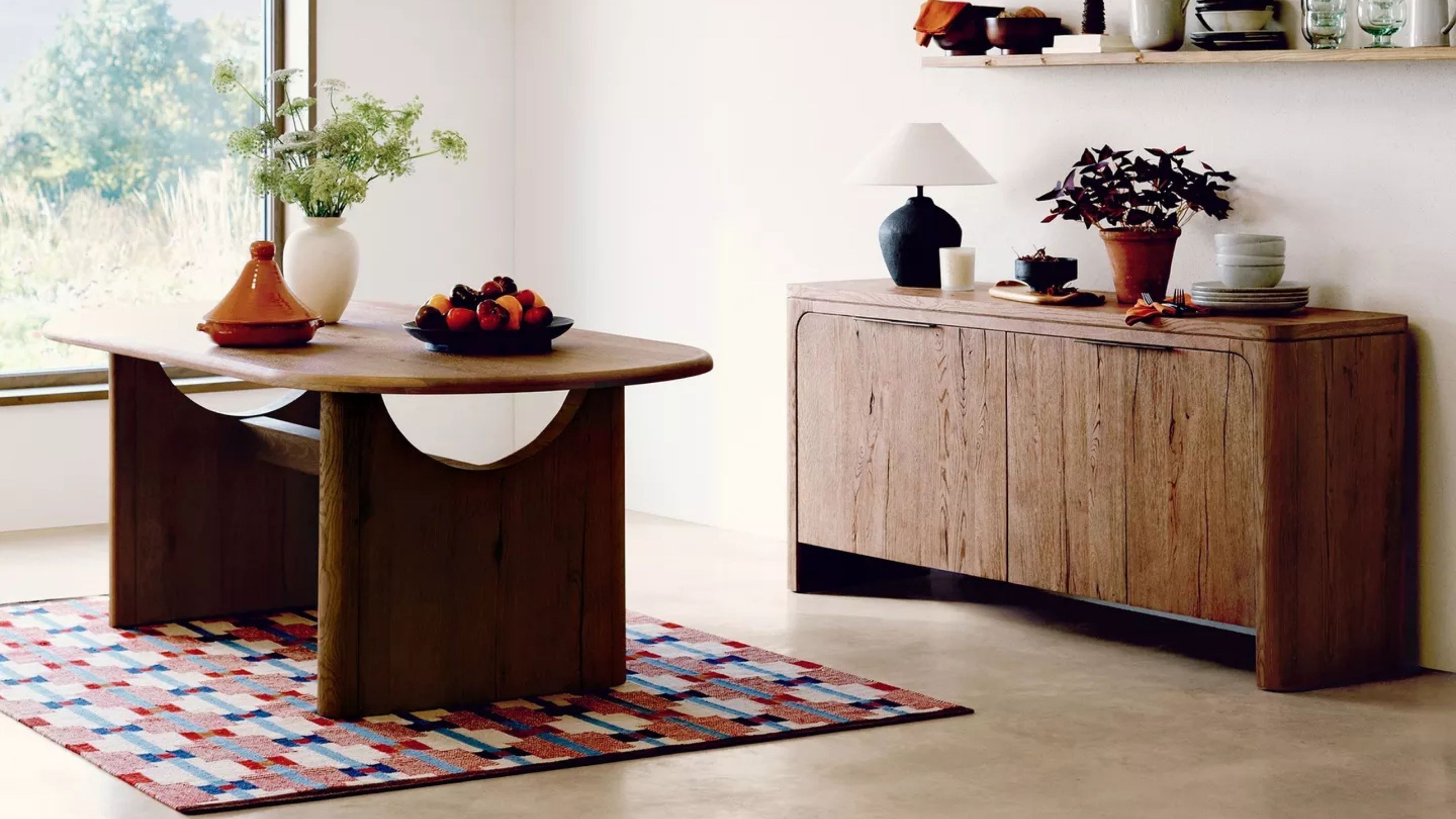 Image of a blue and red modern geometric rug under a wooden dining table in a white room with concrete floors. 
