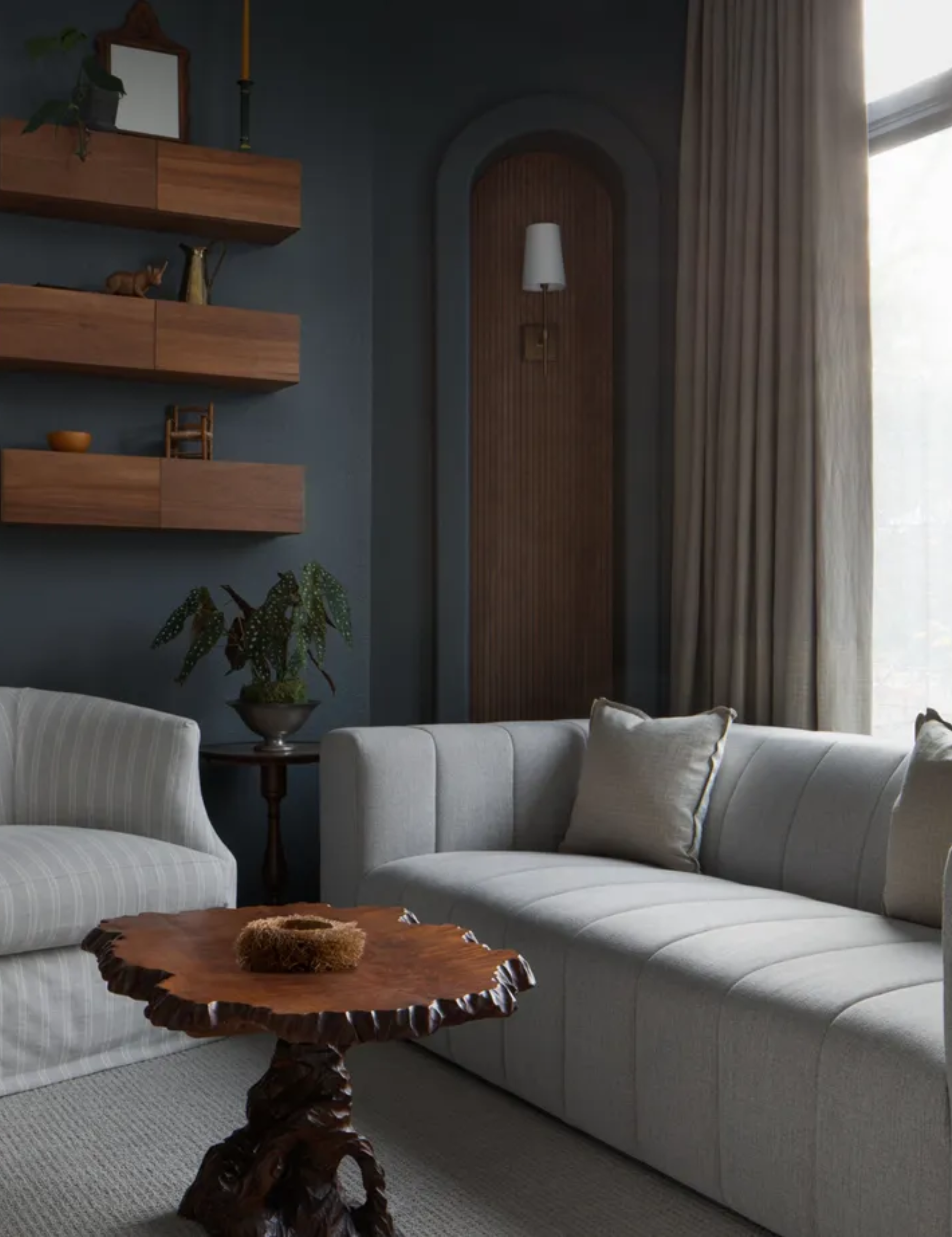 dark blue painted bedroom with a large window and a seating area created below with a small grey sofa, a striped loose linen armchair and a vintage coffee table