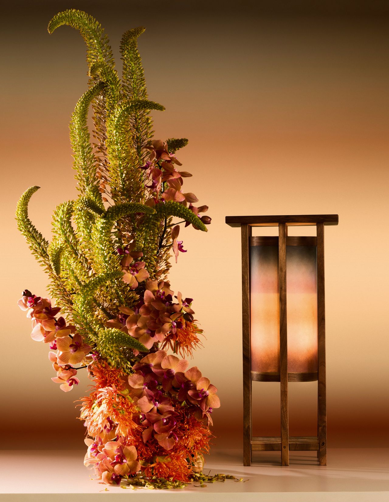 Image of an artisanal lantern lamp with an ombre shade next to an artisanal foliage structure in a display setup that is drenched in a matching ombre background.