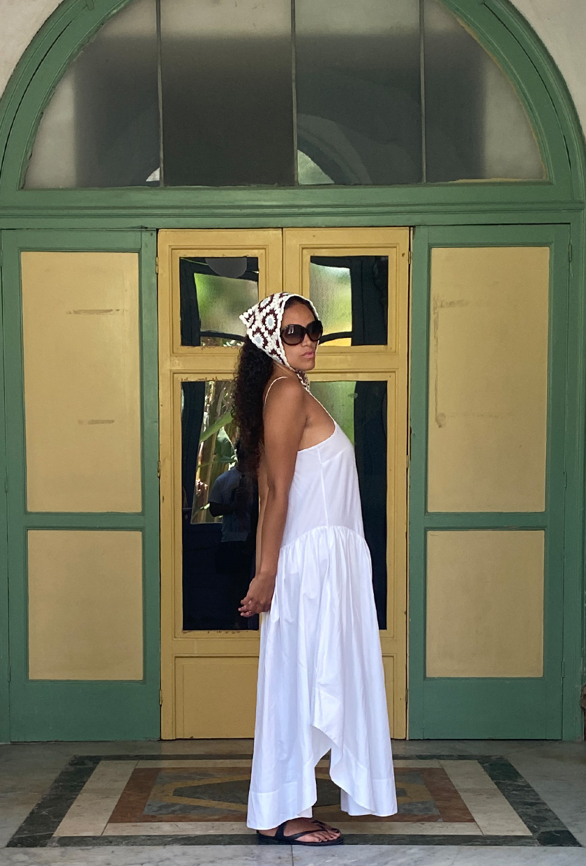 How to pack for Italy is shown in a picture of a fashion editor wearing a white dress with black sandals, a crochet kerchief headscarf, and oversized sunglasses
