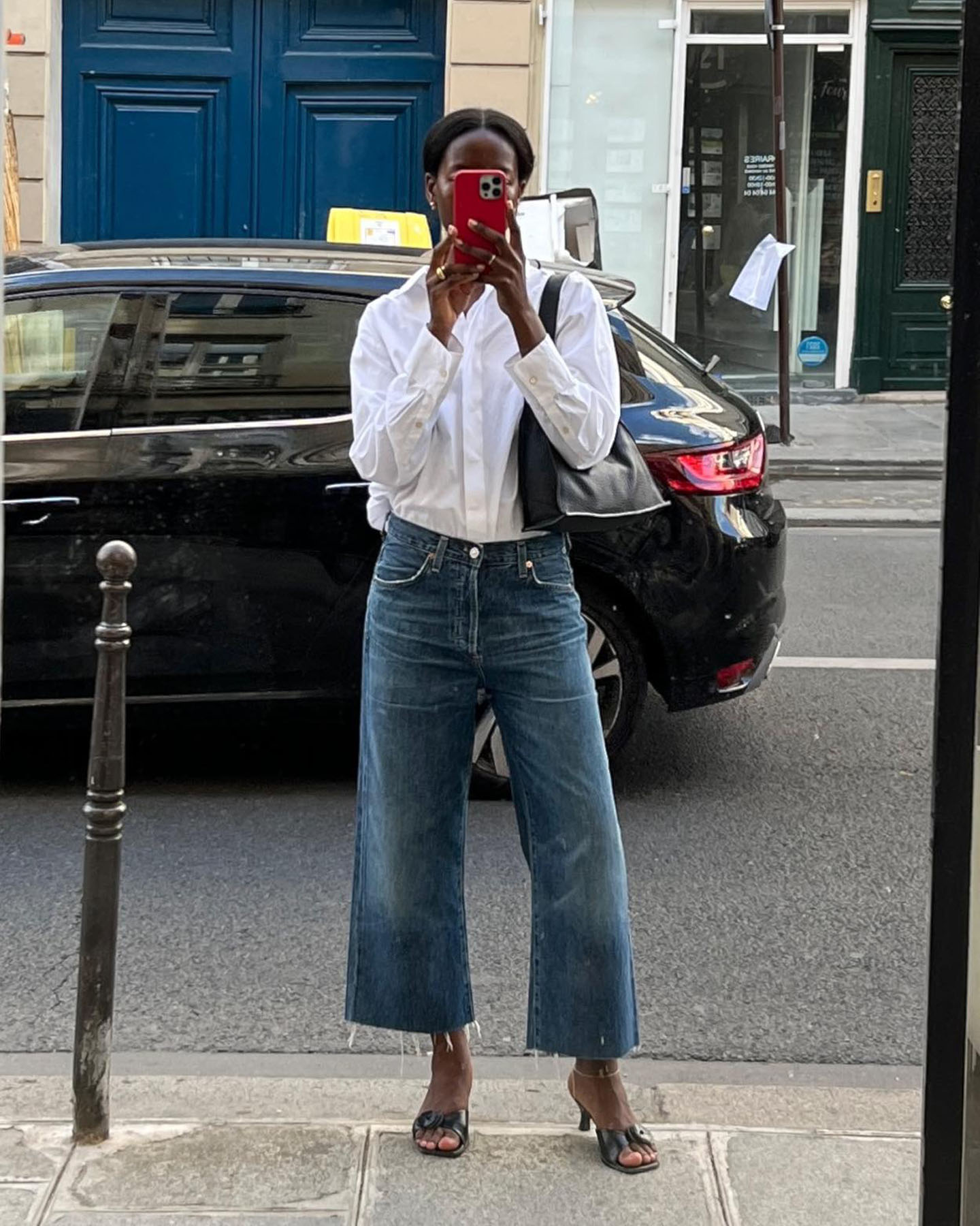 fashion influencer Sylvie Mus takes a mirror selfie on the streets of Paris wearing a white button-down shirt, cropped wideleg raw-hem jeans, and mule sandals