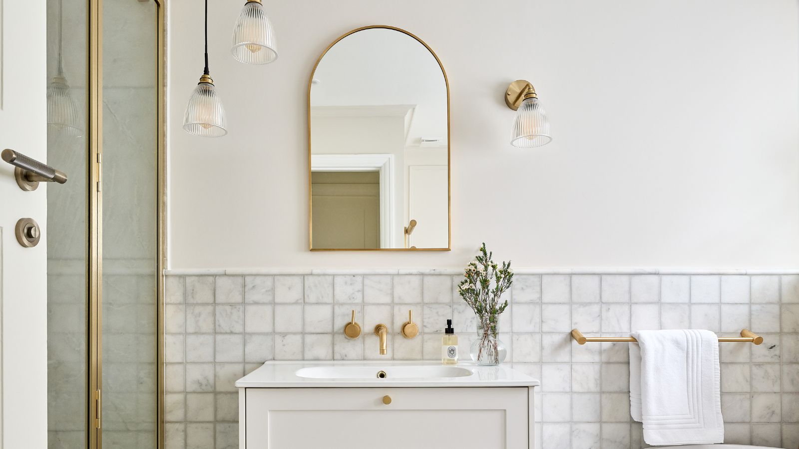 A white bathroom with a gold shower and mirror, marble tiles, and a white sink vanity with gold handles and a gold tap above it