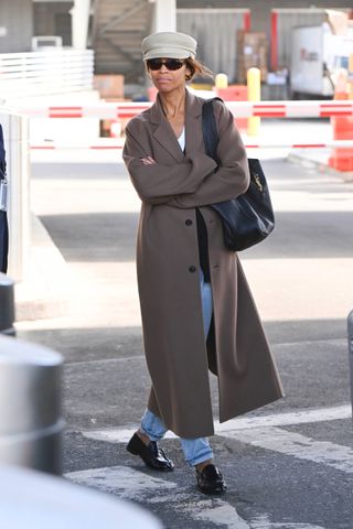 Zoe Saldana is seen at John F. Kennedy International Airport in New York City on October 14, 2024. She is wearing a hat, a brown coat, a black cardigan, jeans and black Saint Laurent loafers. She carries a Saint Laurent shoulder bag.