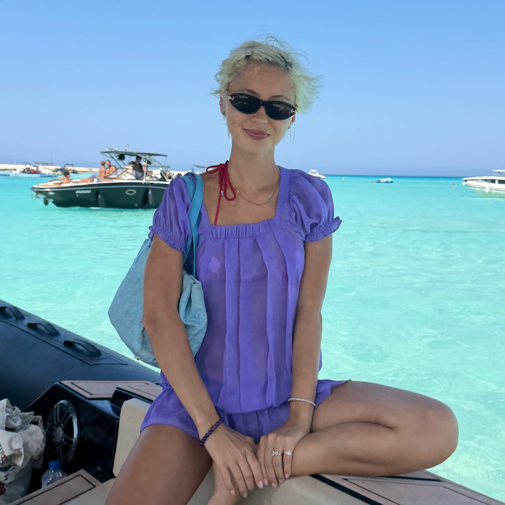 Iris Law sits on a boat wearing a purple minidress with a turquoise bag.