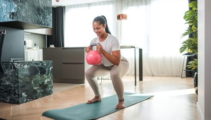 Woman performs goblet squat at home holding pink kettlebell. She is standing barefoot on a green exercise mat, wearing beige athletic leggings and a white short sleeve top.