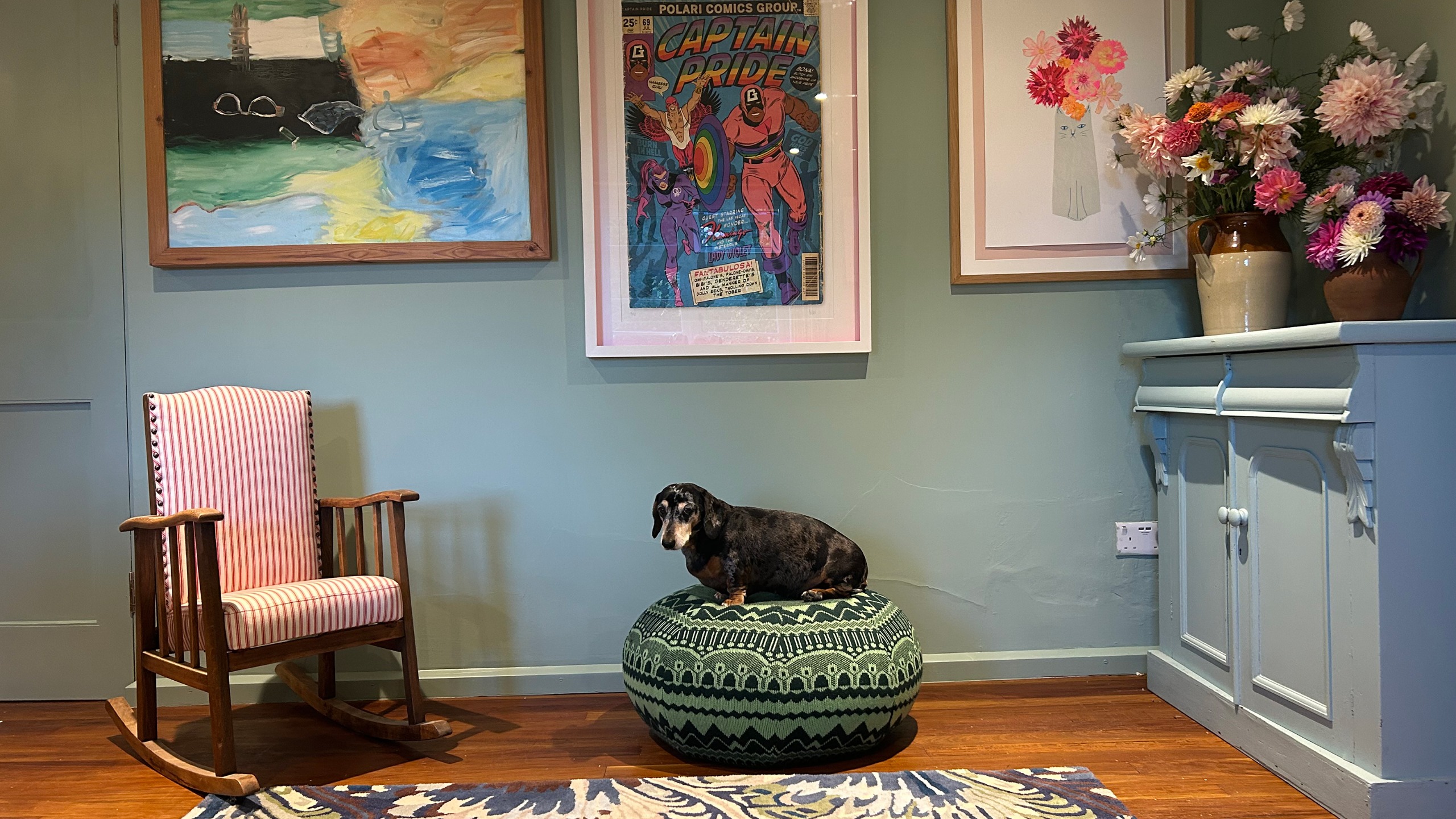 Dog sitting on patterned green pouffe by rocking chair and green wall with framed artworks