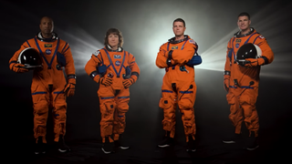 four astronauts standing in a row in orange flight suits. behind them is a dramatic spotlight and darkness