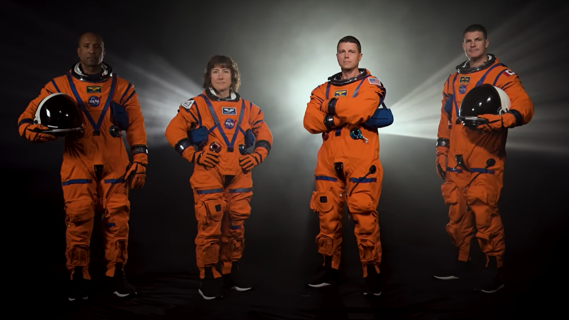 four astronauts standing in a row with a dramatic spotlight behind them. the astronauts wear orange flight suits