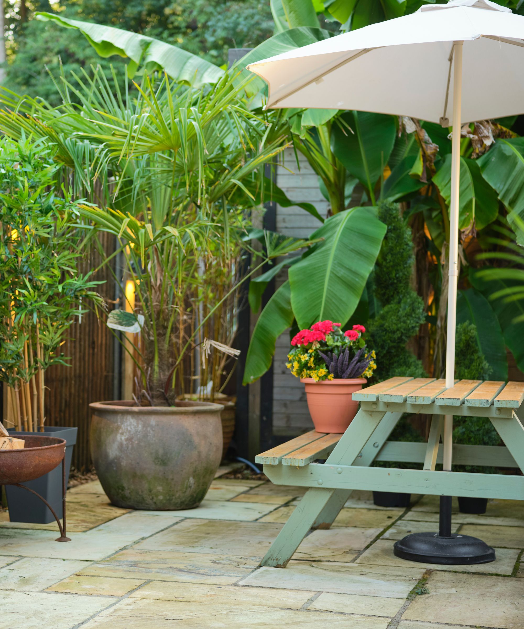 Garden area with green table and bench, large parasol and plants and greenery surrounding the area. On tiled floors in daylight.