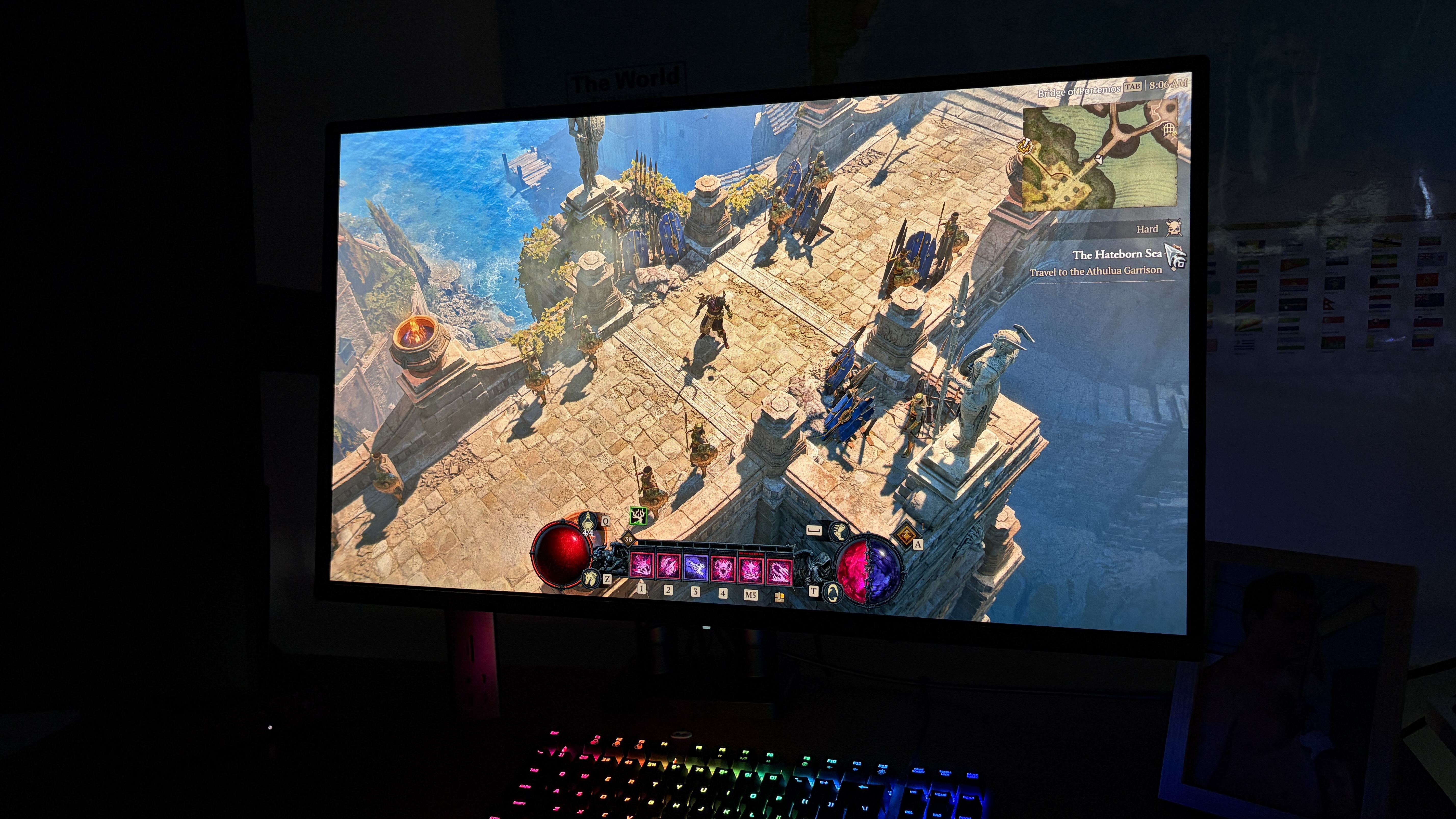 A computer monitor displays a fantasy RPG game set in a vibrant, coastal fortress. Characters stand on stone paths overlooking the sea. A colorful keyboard is visible below.
