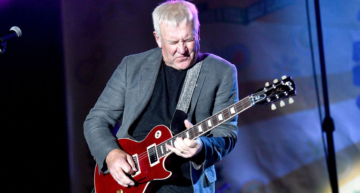 Former Rush guitarist Alex Lifeson hits a note and feels it as he plays a red Gibson Les Paul onstage. He wears a grey blazer and a black T-shirt.