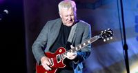 Former Rush guitarist Alex Lifeson hits a note and feels it as he plays a red Gibson Les Paul onstage. He wears a grey blazer and a black T-shirt.