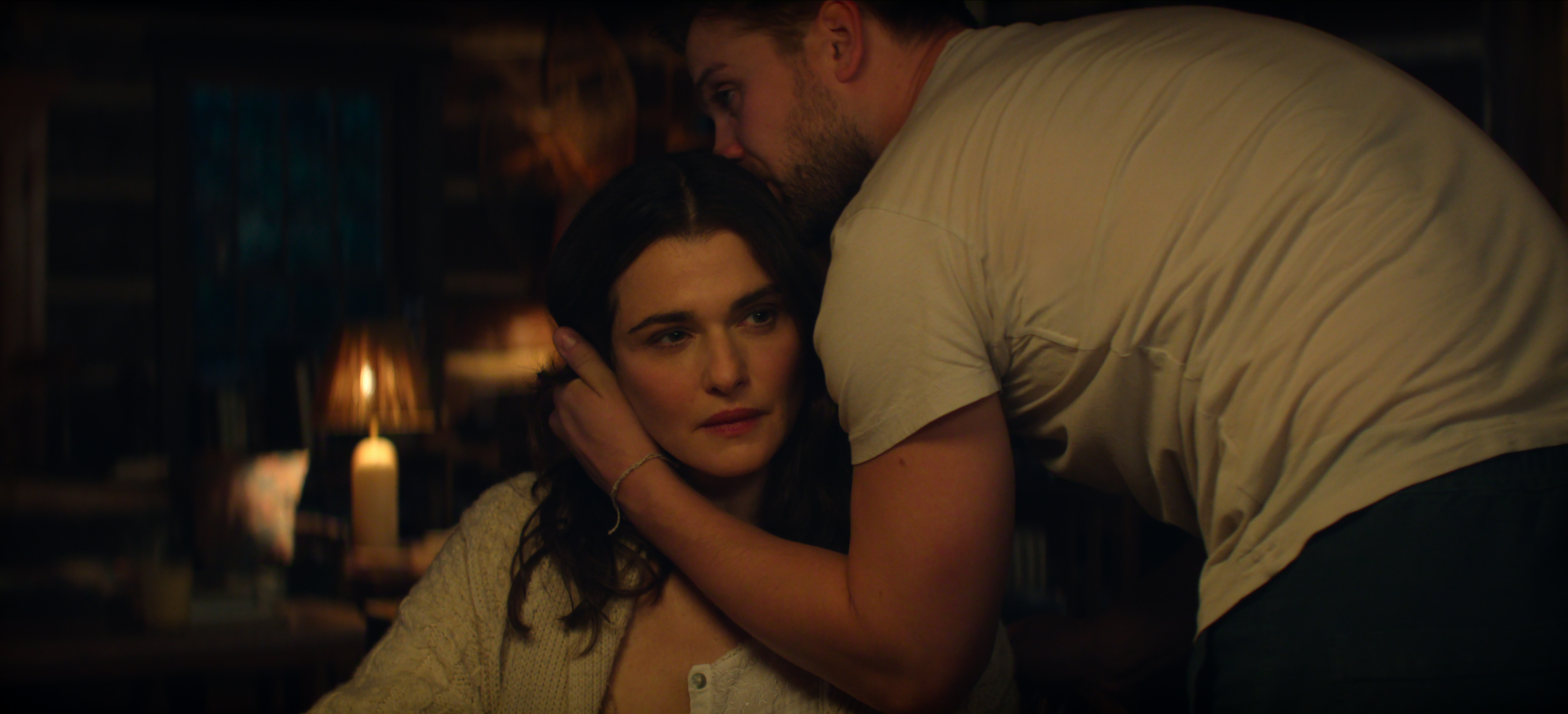 Rachel Weisz as The Protagonist sitting in a chair in a cabin as Leo Woodall as Vladimir stands and wraps his arm around her neck in Episode 108 of Vladimir