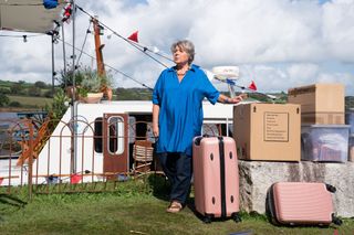 Anne Lloyd (Barbara Flynn) stands in front of Humphrey and Martha's houseboat, with a pile of boxes and suitcases next to her