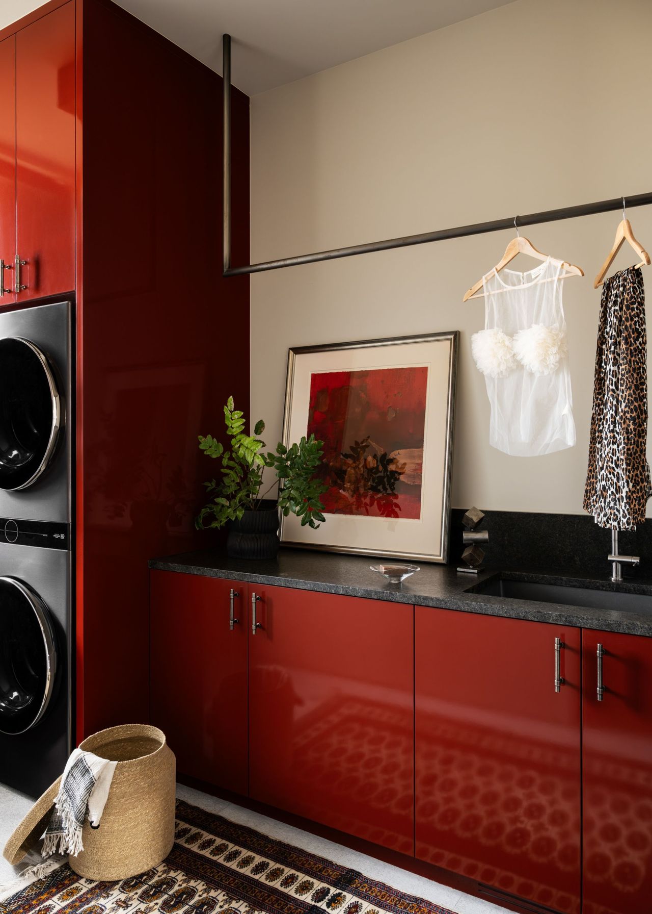 A laundry room with red lacquered cabinets and anthracite counters with a red framed artwork on the counter with a pole for hanging laundry sitting just in front next to the washing machines