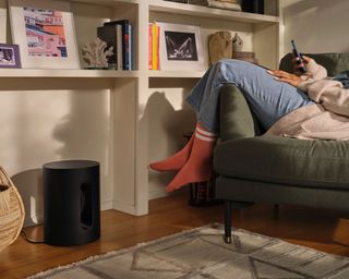 Sonos Sub Mini on the floor beside woman relaxing and two Sonos One SL speakers on the bookshelf