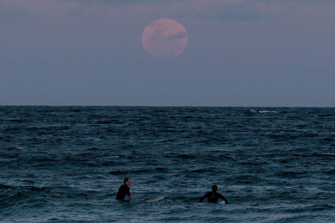 red moon appearing above the sea with surfers in the foreground.