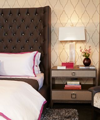 A bedroom with a brown bed with tall winged buttoned headboard, white bedding with fuschia pink piping detail and a brown two-tiered nightstand with a white lamp and pink books and trinkets for accent