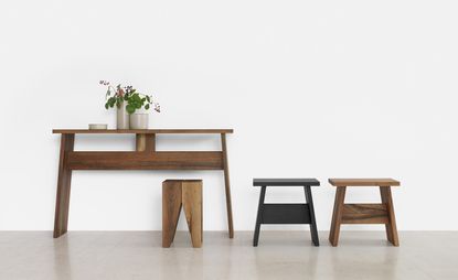 A wooden table is to the left, with a vase with flowers in it. Two stools are to the right of the table, one is black and the other one is plain wood.