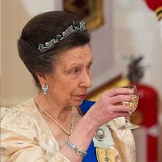 Princess Anne holds up a glass while wearing a cream dress and a small tiara