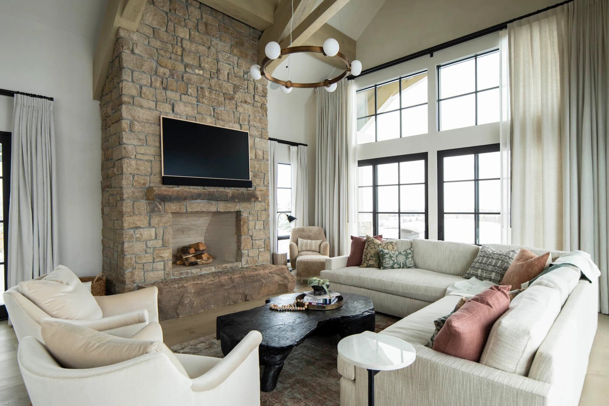 living room with stone fireplace and a TV mounted above, wall of windows, white L-shaped sofa with two white armchairs opposite, and a black timber coffee table