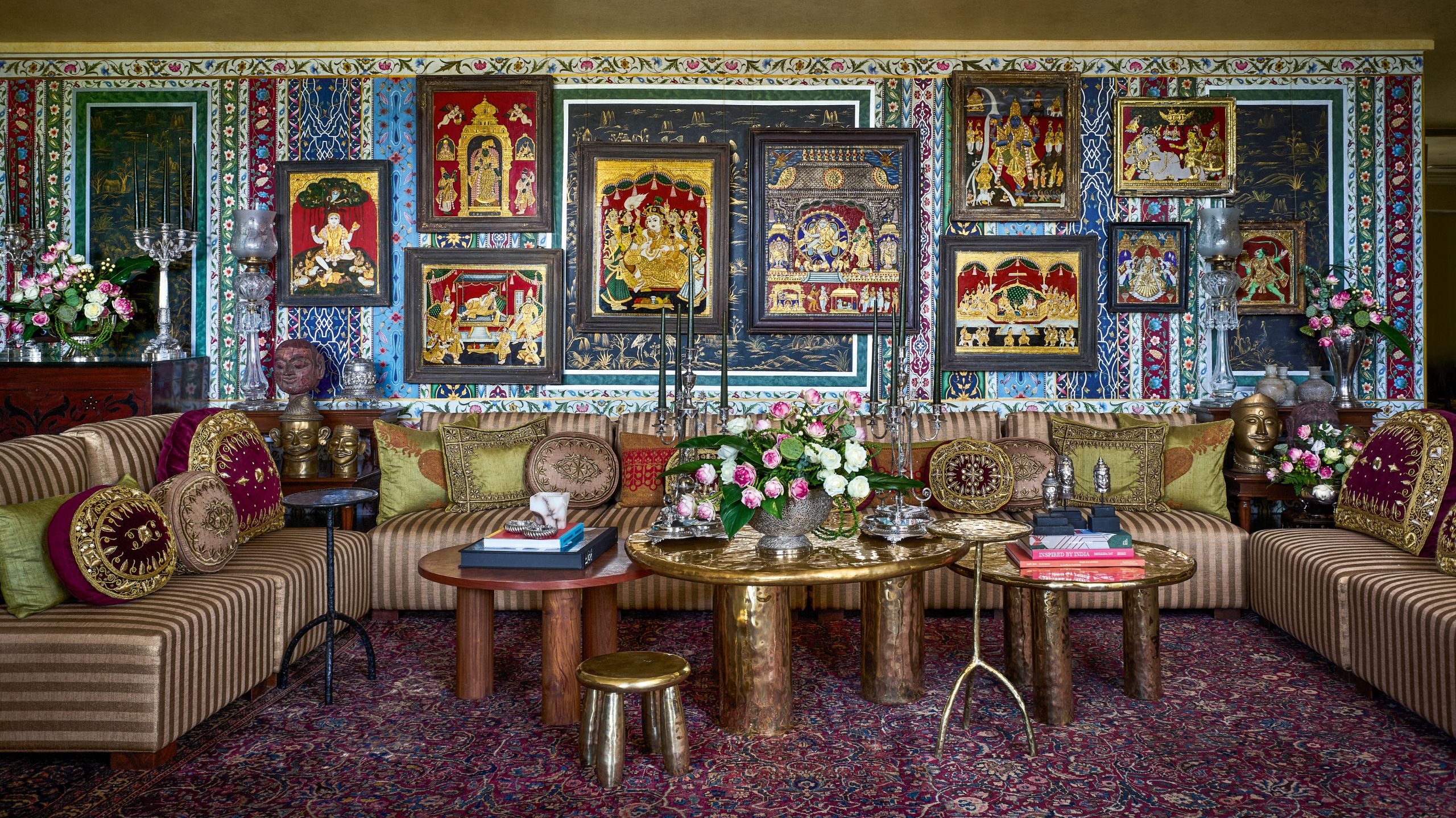 Opulent living room featuring a striped sectional sofa, brass coffee tables, and a gallery wall of ornate framed artwork set against vibrant patterned wallpaper.