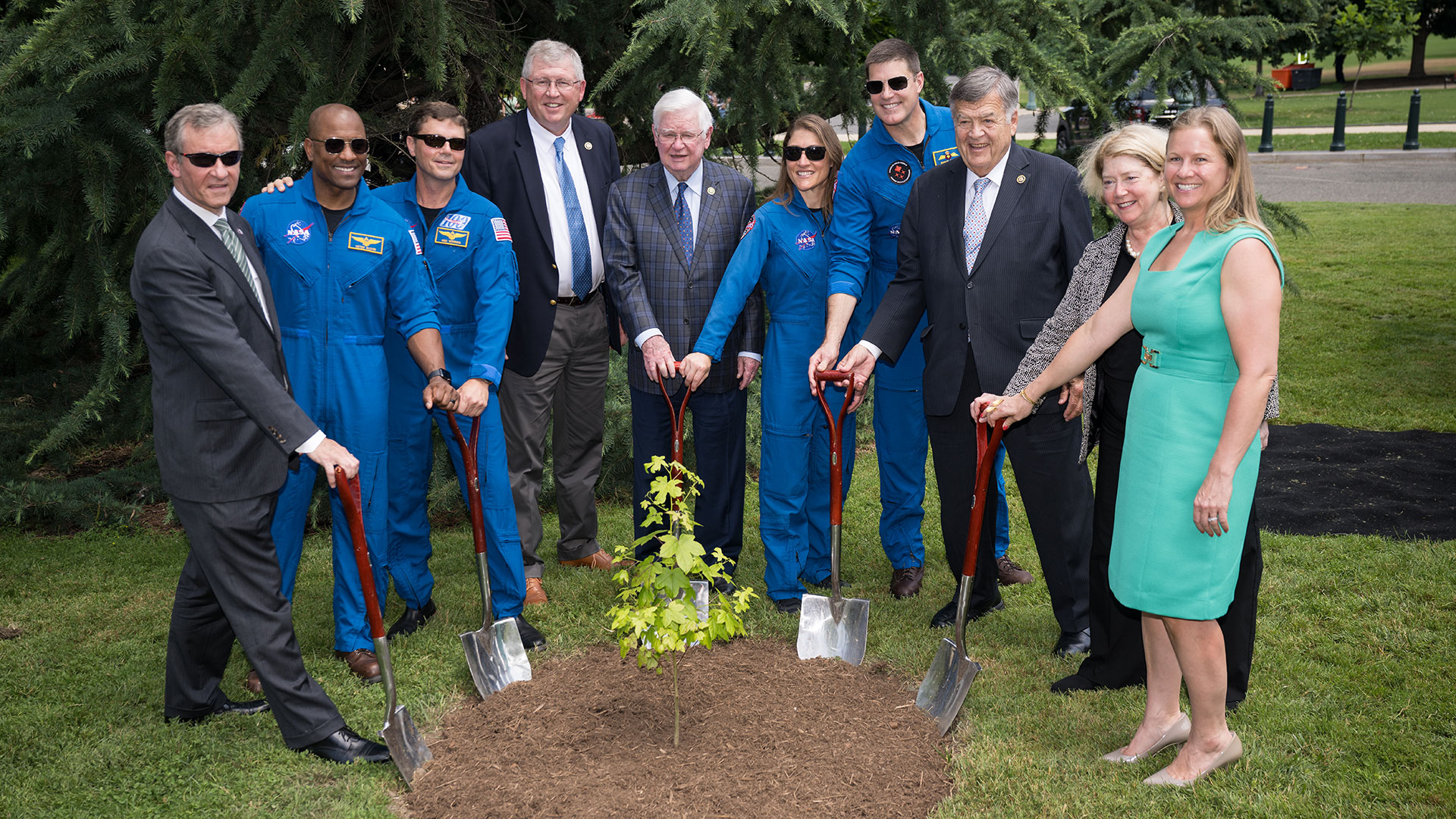 a group of people in suits and blue flight suits smile while planting a tiny tree