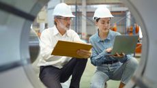 An older engineer in a white collared shirt examines a laptop held by a young colleague. The camera is looking at them trhough an industrial tube.