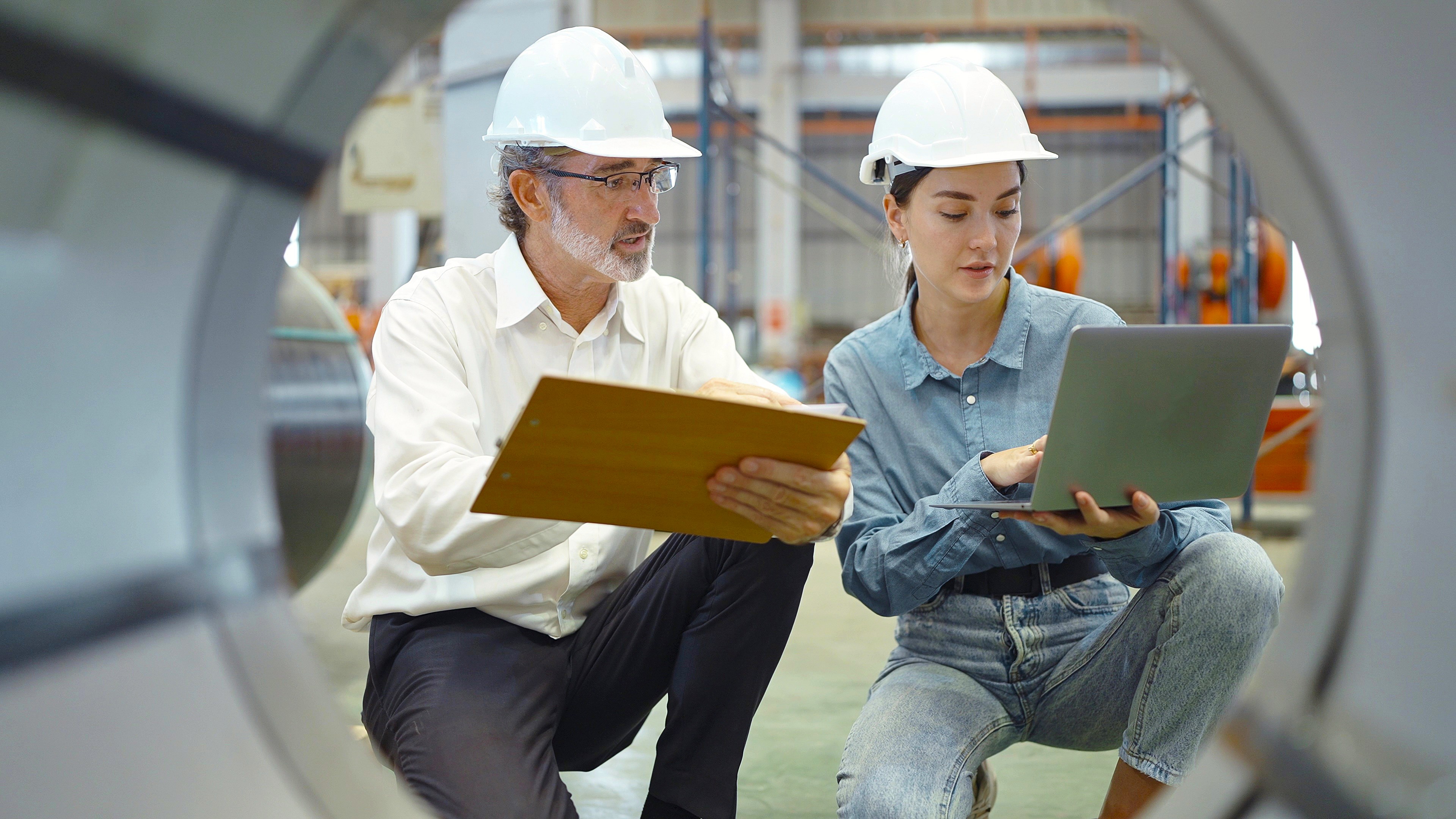 An older engineer in a white collared shirt examines a laptop held by a young colleague. The camera is looking at them trhough an industrial tube.
