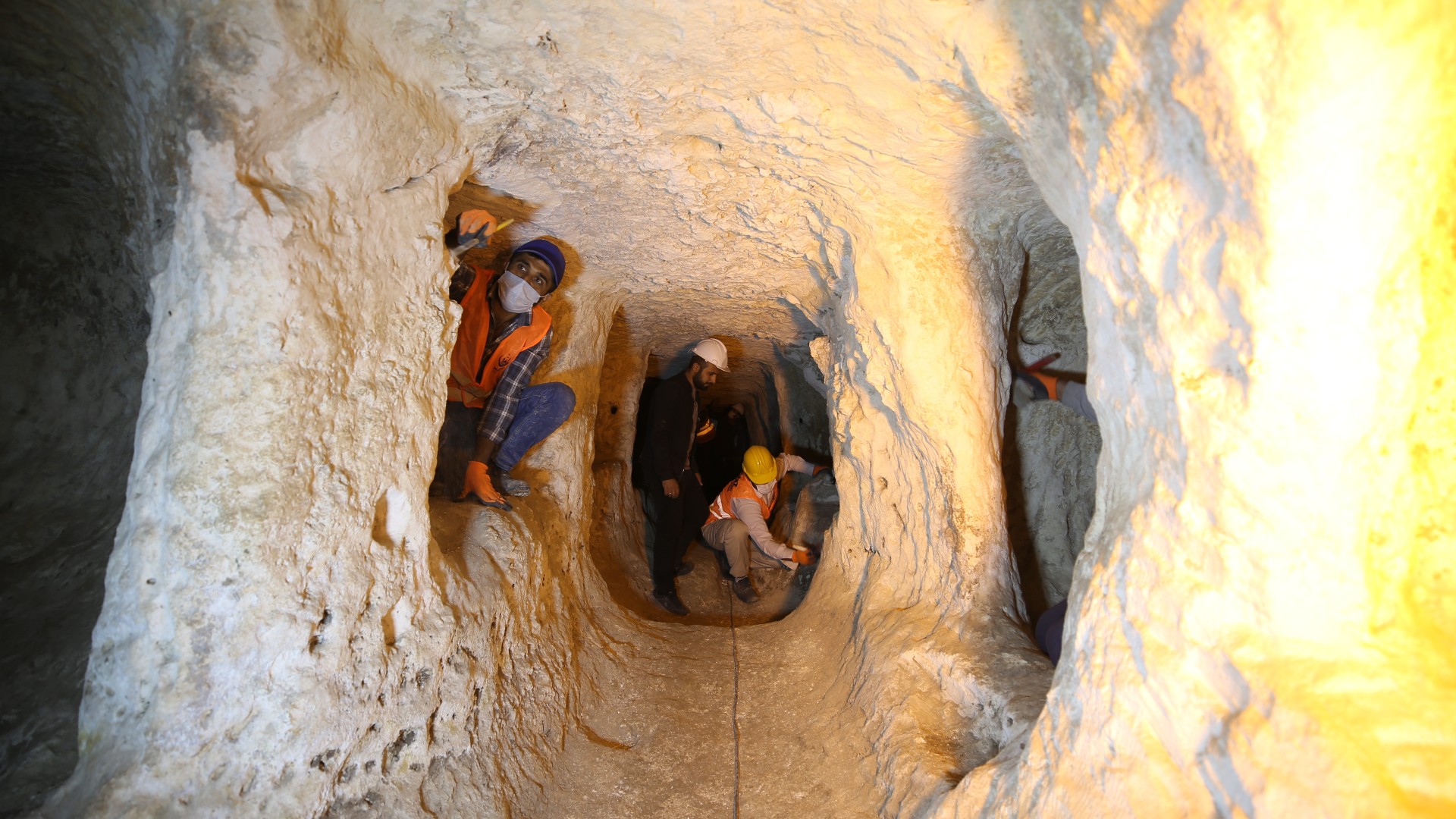 A team of 3 people who are wearing hard hats, masks and high visibility safety vests are exploring a brightly lit tunnel of an underground cave thought to be a city.