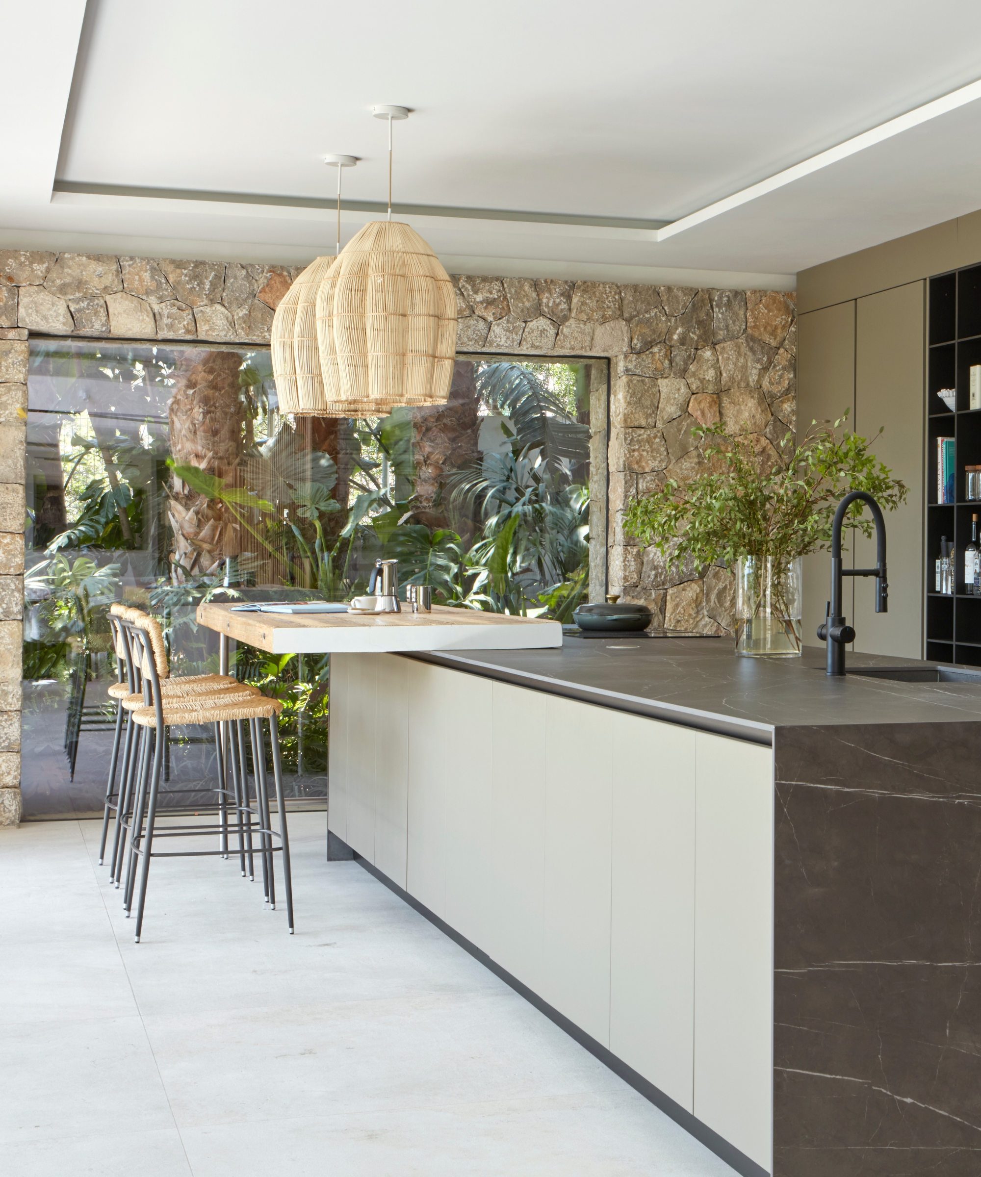 A Tuscan-inspired kitchen with exposed stone walls, woven pendant lights, pale green cabinets, and dark stone countertops
