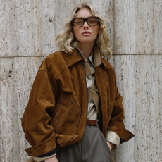 Model Elsa Hosk wearing suede jacket sunglasses and gray pants standing against stone background.