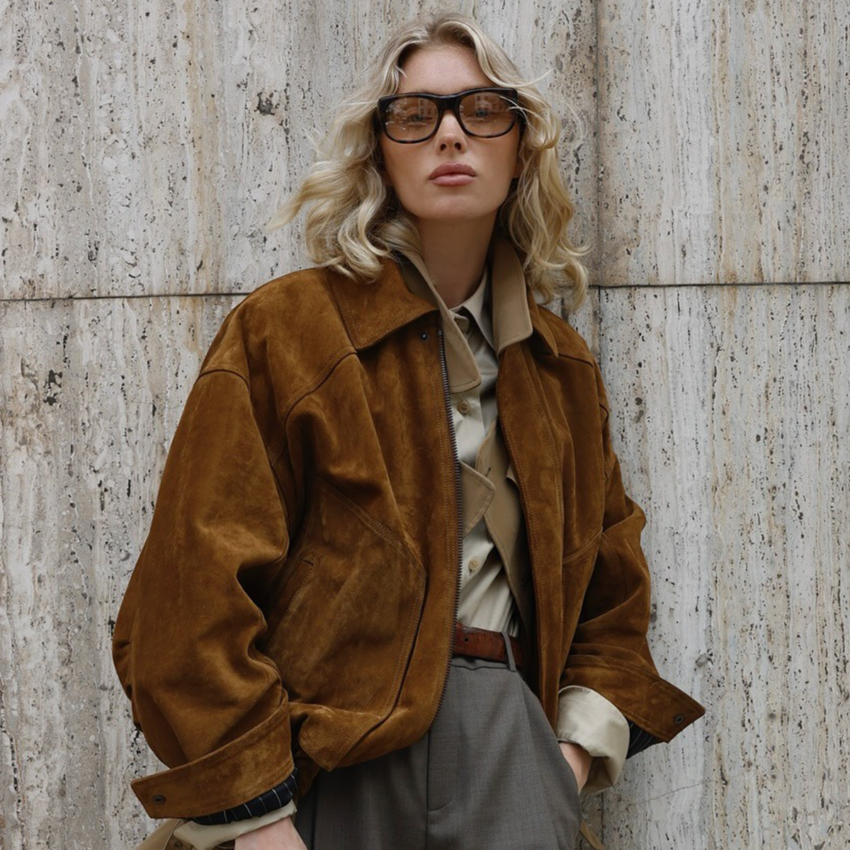 Model Elsa Hosk wearing suede jacket sunglasses and gray pants standing against stone background.