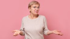 An older woman shrugs while standing against a pink background.
