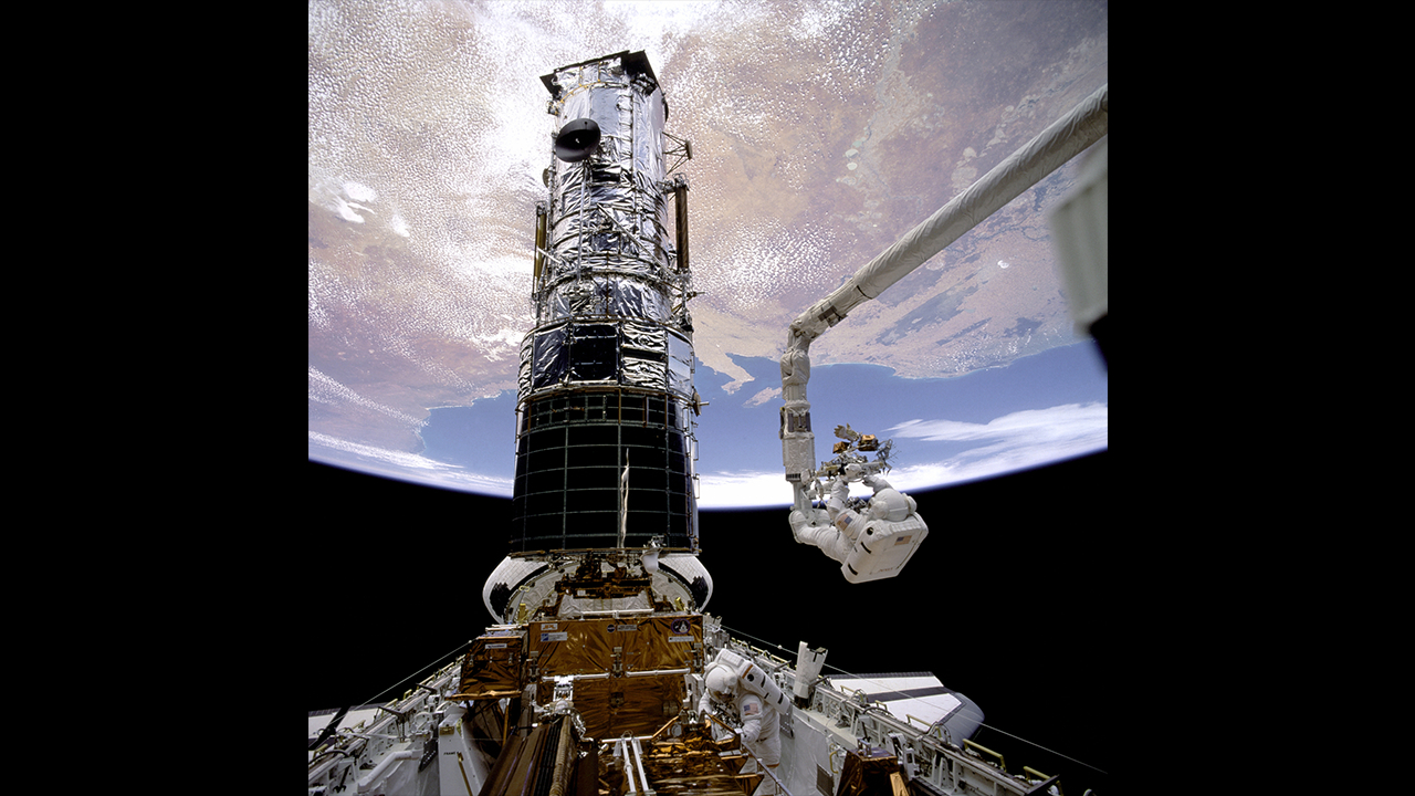 a large chrom cylinder (Hubble) is attachted to the payload bay of a space shuttle. Earth is seen hanging above, a mechanical arem extends on the right.