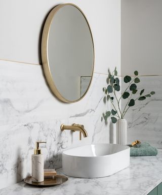 A corner of a bathroom with white walls, marble tiles, a circular gold mirror hanging on the wall, a white sink with a gold tap, a golden tray with a white soap dispenser, and a white vase with plant stems