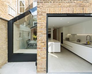 A glass small house extension by Yard Architects attached to a brick terraced property, with a modern, all-white kitchen inside and a large white island