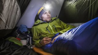 A man in his sleeping bag in a tent wearing a head torch