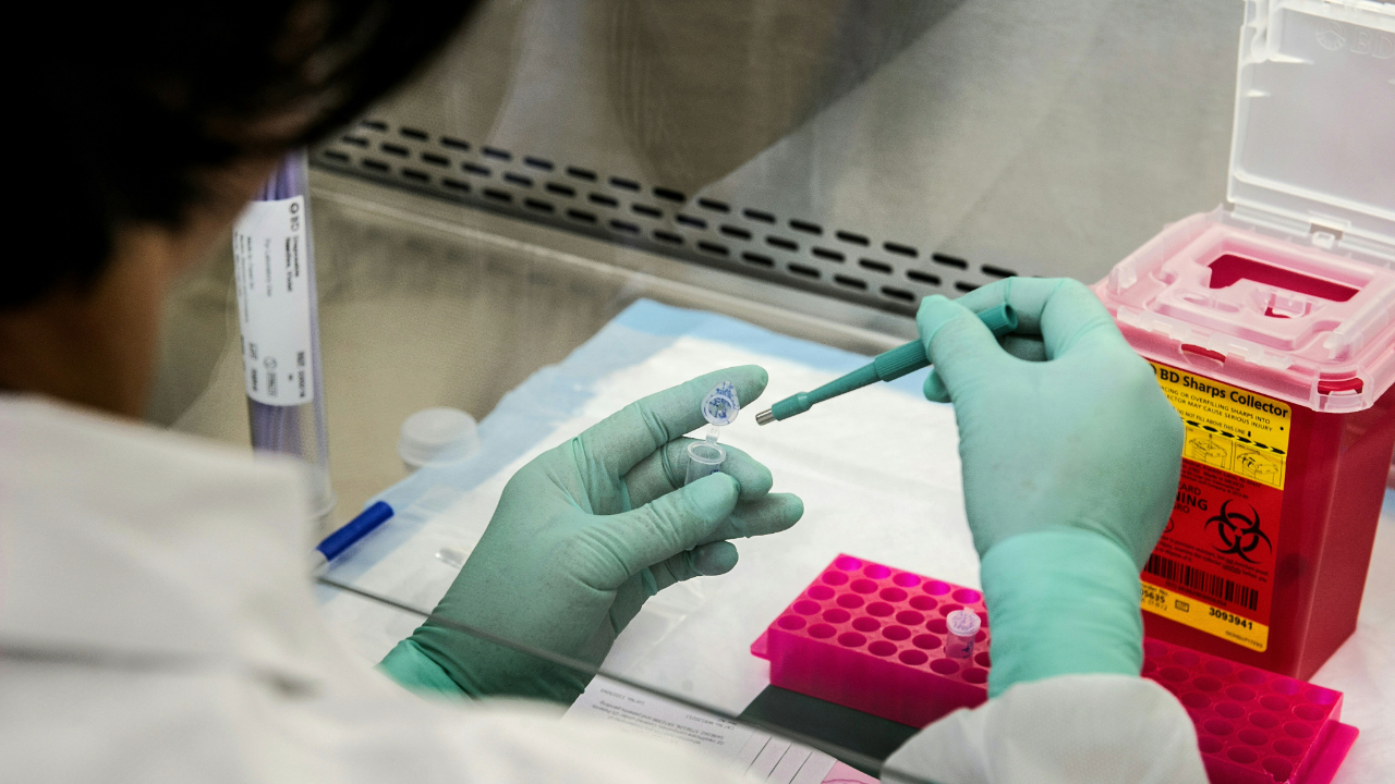 A person wearing light green gloves works in a clean environment next to a sharps container, pipetting liquid into a small plastic capsule
